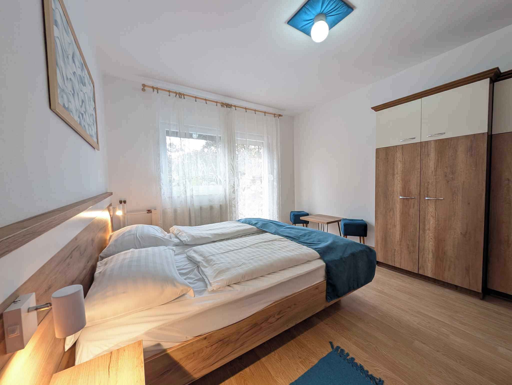 A bedroom with a bed, bedside lamps, a window with sheer curtains, a desk with two chairs, and a wardrobe.