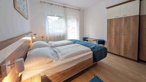 Ein Schlafzimmer mit Bett, Nachttischlampen, einem Fenster mit durchsichtigen Vorhängen, einem Schreibtisch mit zwei Stühlen und einem Kleiderschrank.