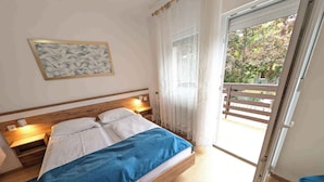 Ein Schlafzimmer mit einem hölzernen Bett, weißer Bettwäsche, einer blauen Decke, einem Nachttisch mit Lampe, einem Wandbild und einer Glastür, die auf einen Balkon im Freien führt.