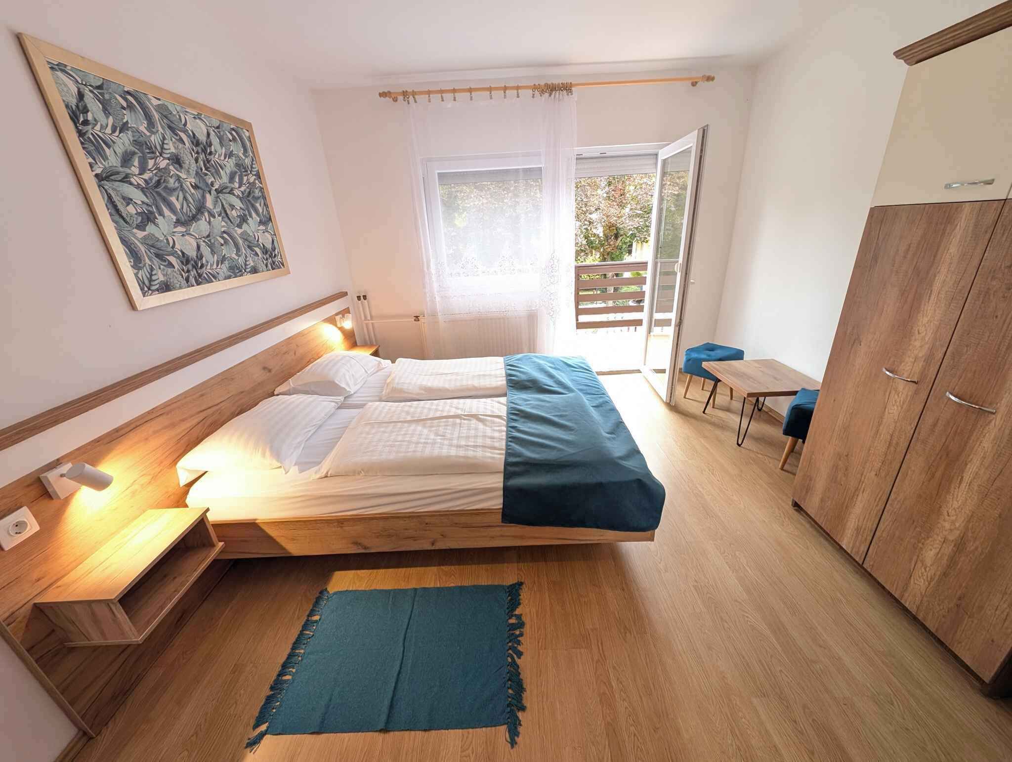 A bedroom with a bed, bedside table, lamp, wardrobe, and a view of the outside.