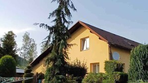 Une maison à deux étages, dotée d’un toit en pente, entourée d’un jardin bien entretenu et d’arbres.