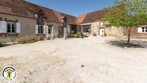 Une cour avec un sol de gravier, un petit arbre et un bâtiment à la façade en briques et aux volets blancs.