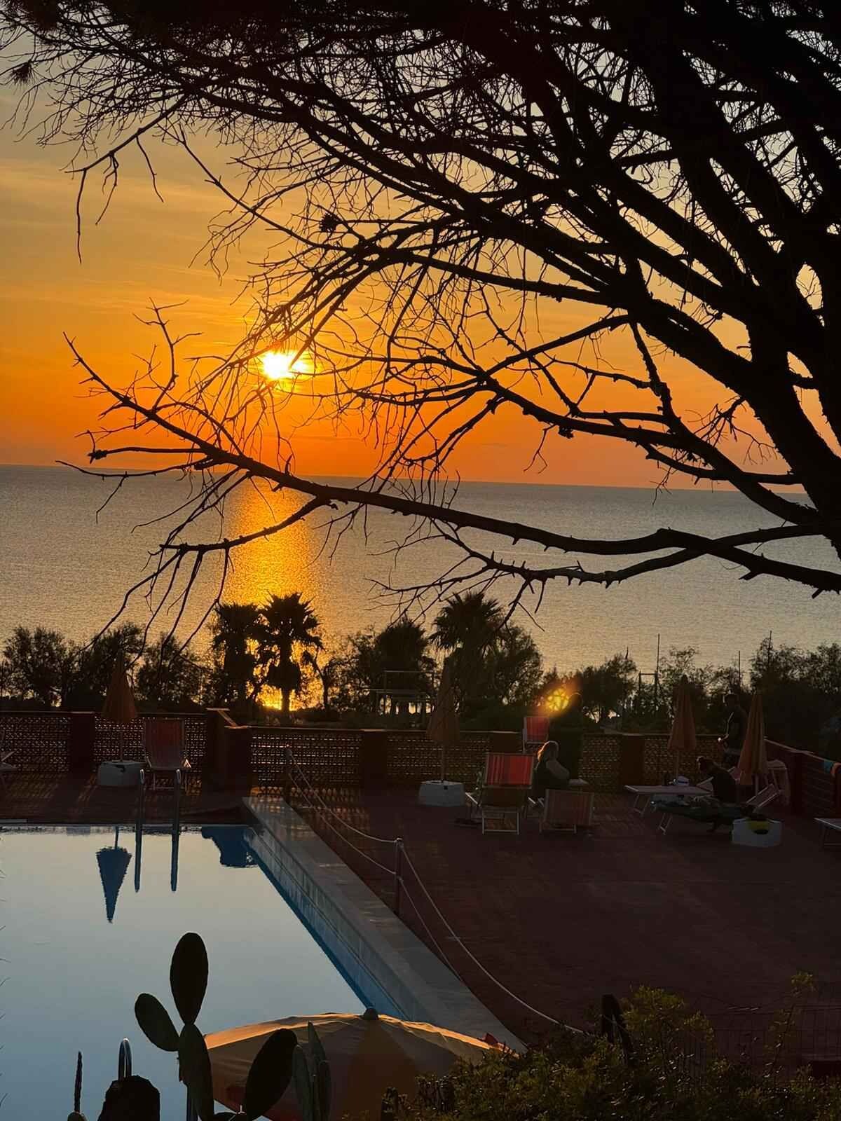 Un espace piscine doté de chaises longues et de parasols, offrant une vue sur le coucher de soleil sur l’océan.