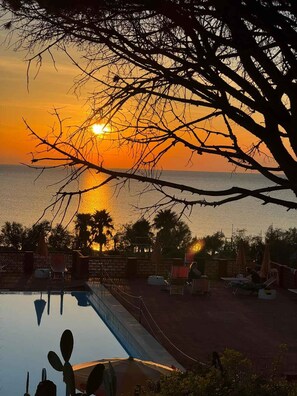 A pool area with lounge chairs and umbrellas, overlooking a sunset over the ocean.
