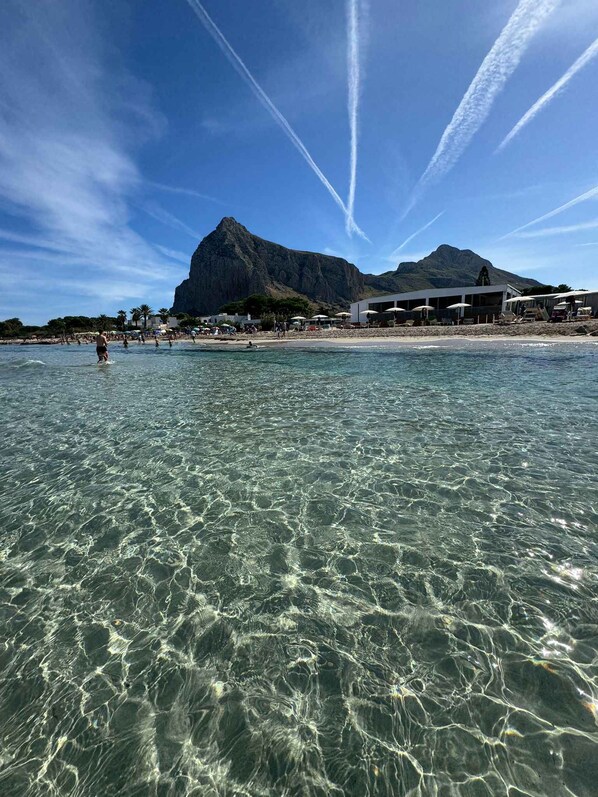 A beach with clear water, a mountain in the background, and a building with multiple levels.