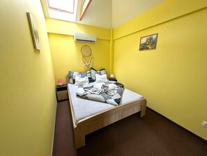 Une petite chambre à coucher aux murs jaune vif, avec un lit simple, une table de chevet, un climatiseur fixé au mur, un attrape-rêves et un tableau encadré.