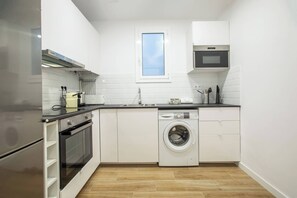 Une cuisine compacte avec des meubles blancs, un four encastré, un four à micro-ondes, un lave-linge et une fenêtre au-dessus de l’évier.