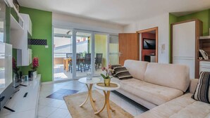 Una sala de estar moderna con un sofá, una mesa de centro y un televisor. Hay un ventanal corredero que da a un área exterior con una mesa y sillas.