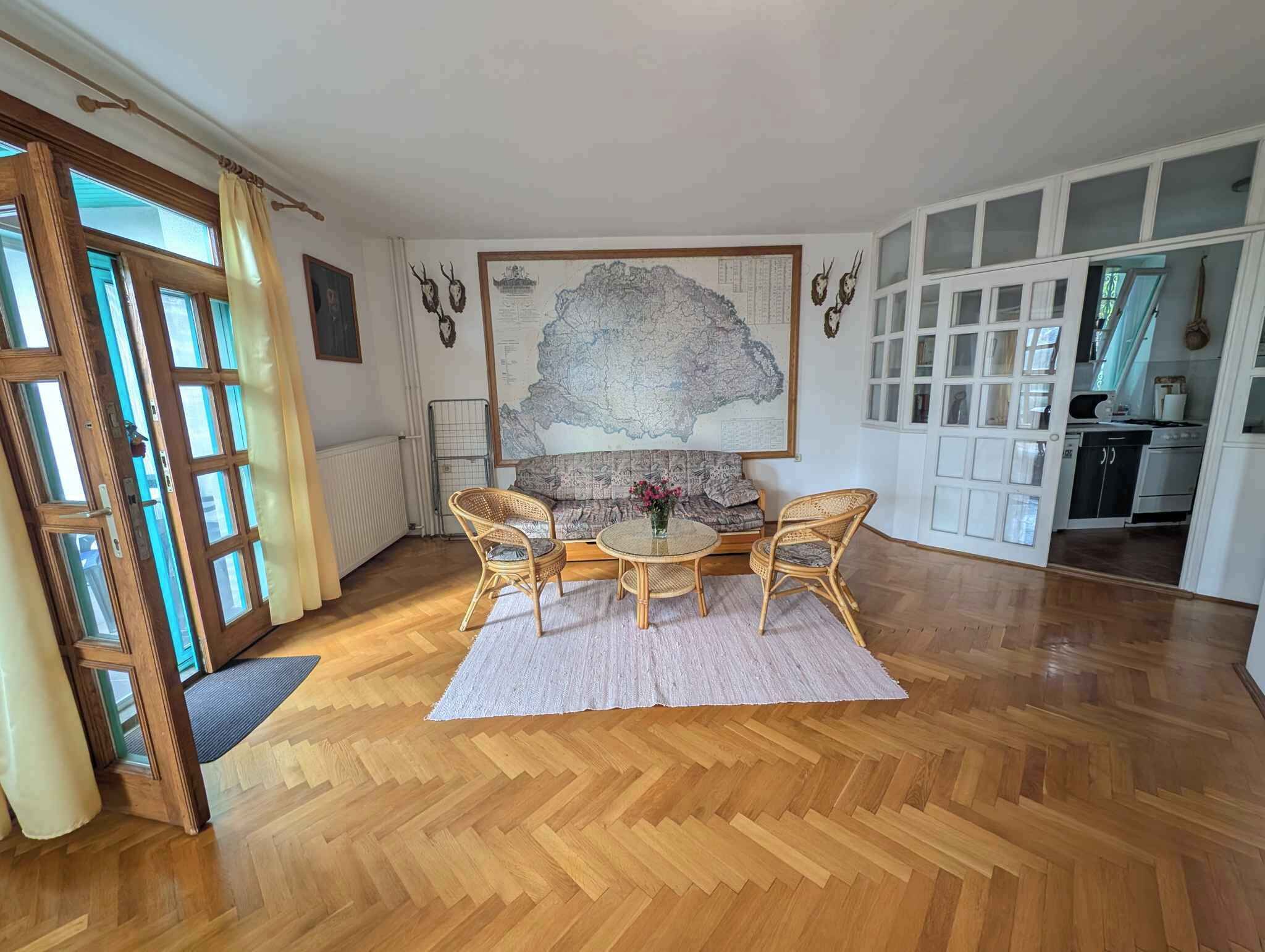 A spacious room with a wooden herringbone floor, a round dining table with four chairs, a large map on the wall, and a kitchen area visible through an open door.