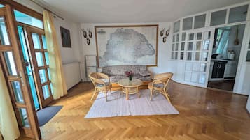 A spacious room with a wooden herringbone floor, a round dining table with four chairs, a large map on the wall, and a kitchen area visible through an open door.