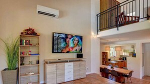 A spacious living area with a dining table, a bookshelf, and a mounted TV displaying a song list.