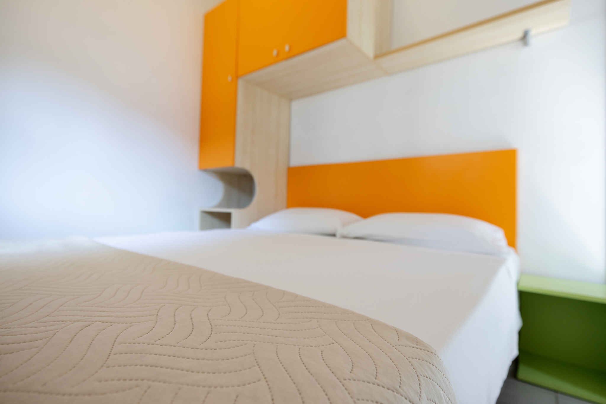 Quarto de hotel com cama, paredes brancas e detalhes em laranja.
