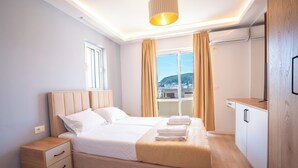 A modern hotel room with a large bed, a bedside table, a wardrobe, and a view of the city. - iLLYRIA HOTEL (Ulcinj)
