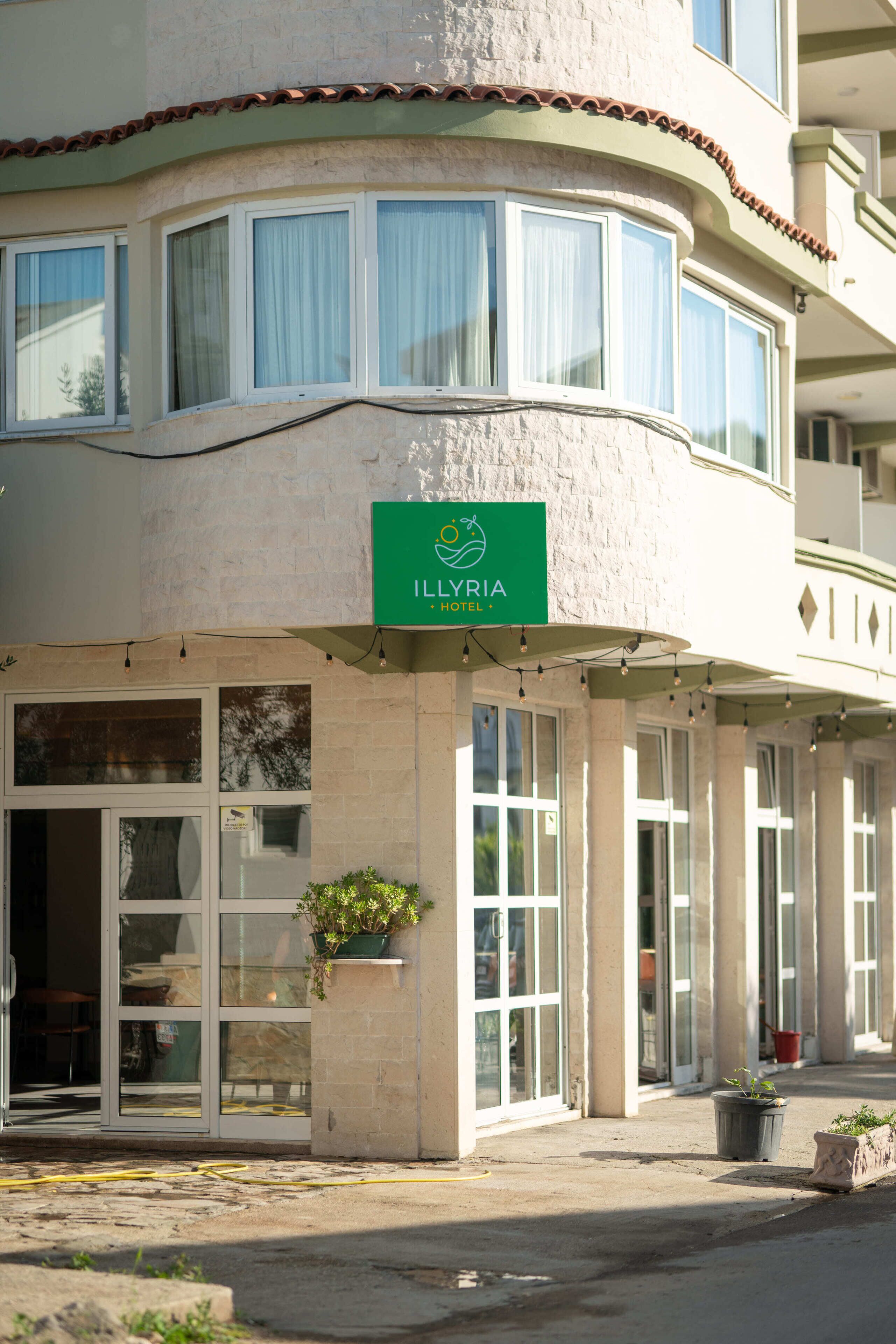 Entrada de un hotel con un letrero que dice "ILLYRIA HOTEL", amplias ventanas y plantas en macetas.