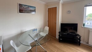 A living room with a glass table, white chairs, a black entertainment unit, a wooden door, a clock, and a window with a view. - Convenient Edinburgh City Centre 2 Rooms + Parking (Edinburgh)