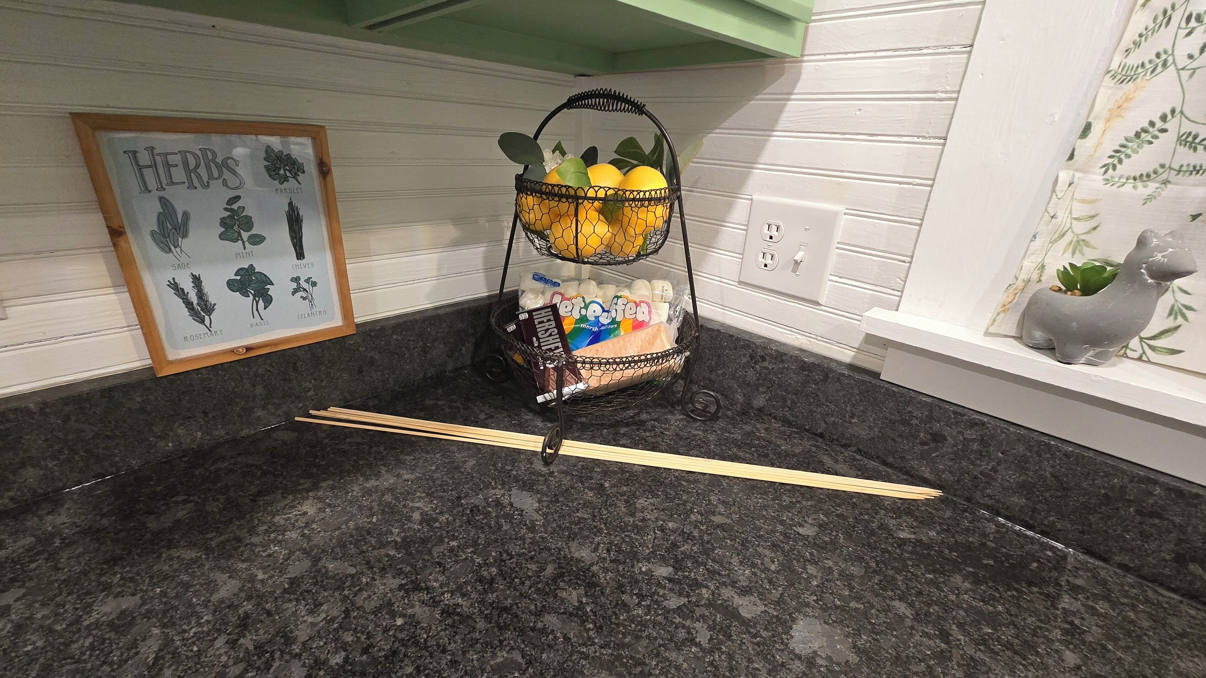 A kitchen counter with a fruit basket, a framed herb illustration, and a small plant.