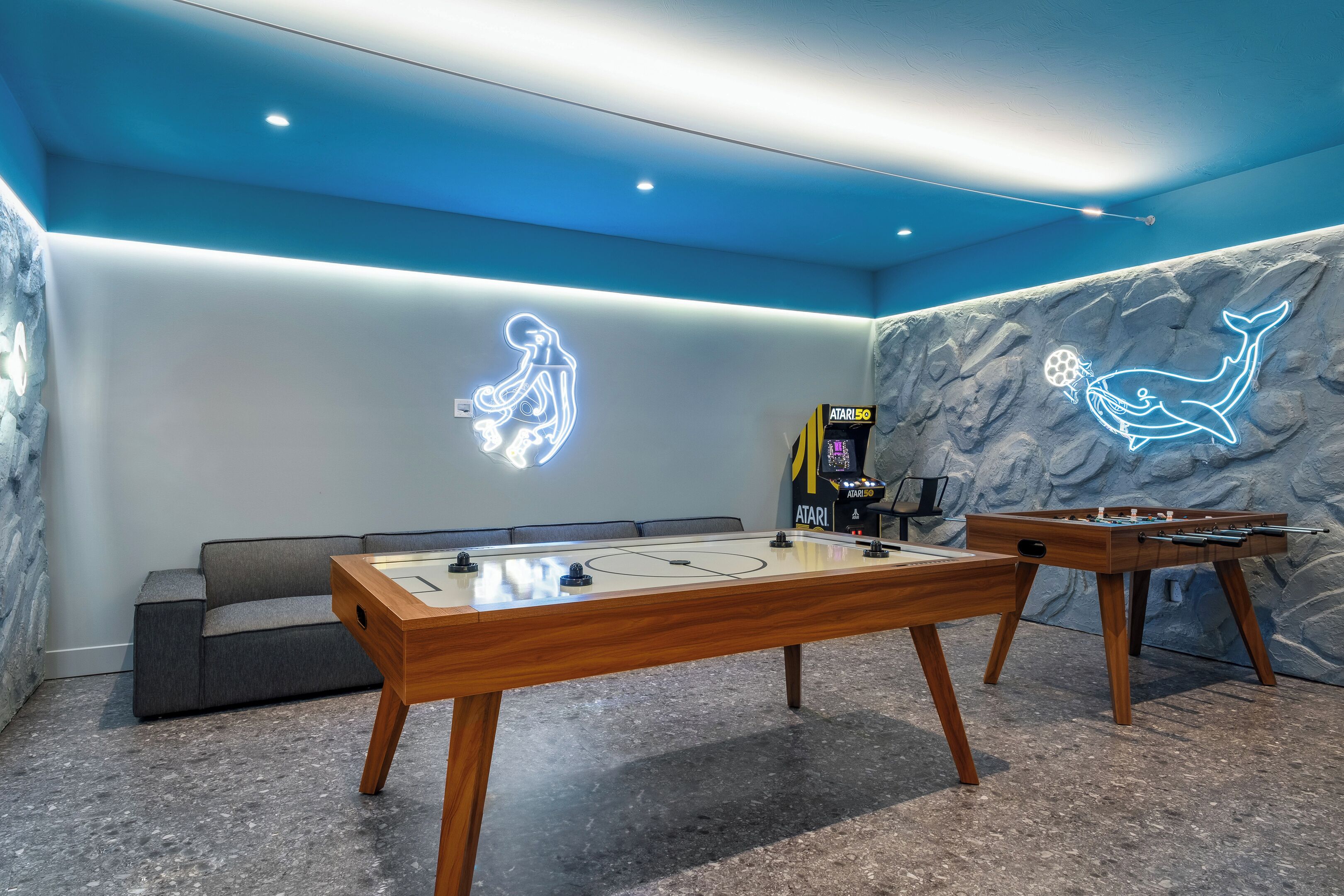 A room with two foosball tables, a sofa, and neon wall art.