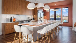 A modern kitchen with wooden cabinets, a large island with bar stools, and a view of the outdoors through large windows. - 7bd 8bath Hot tub, Pool, Game Room, Sauna, Professional Pickleball Court (Fennville)