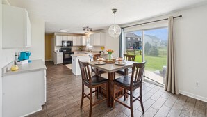Une cuisine moderne avec des meubles blancs, un coin repas avec des chaises en bois et une grande porte-fenêtre coulissante donnant sur un jardin.