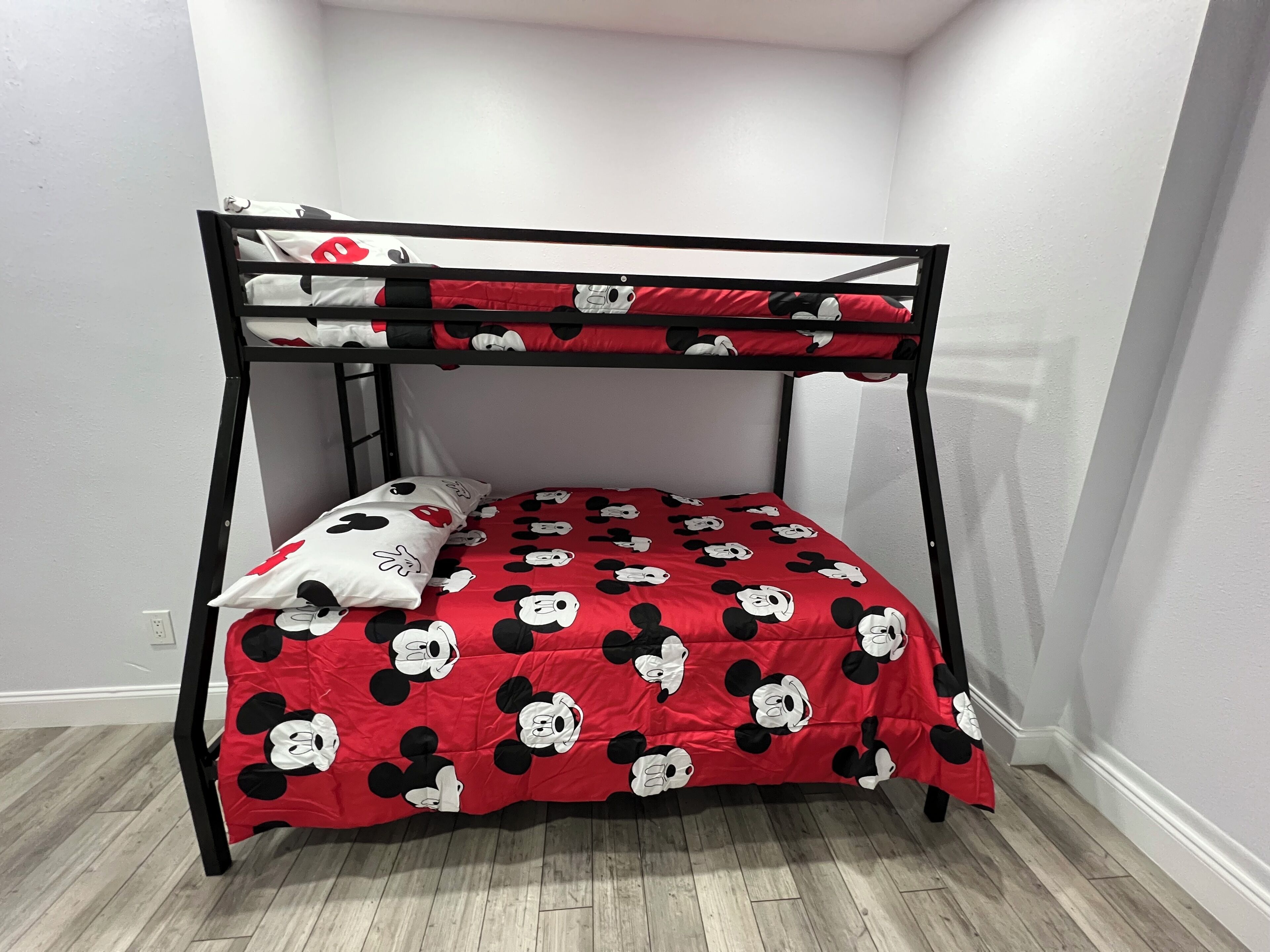 Een stapelbed met een rode en zwarte bedspread met Mickey Mouse thema, een kussen met een vergelijkbaar ontwerp, en een houten vloer.