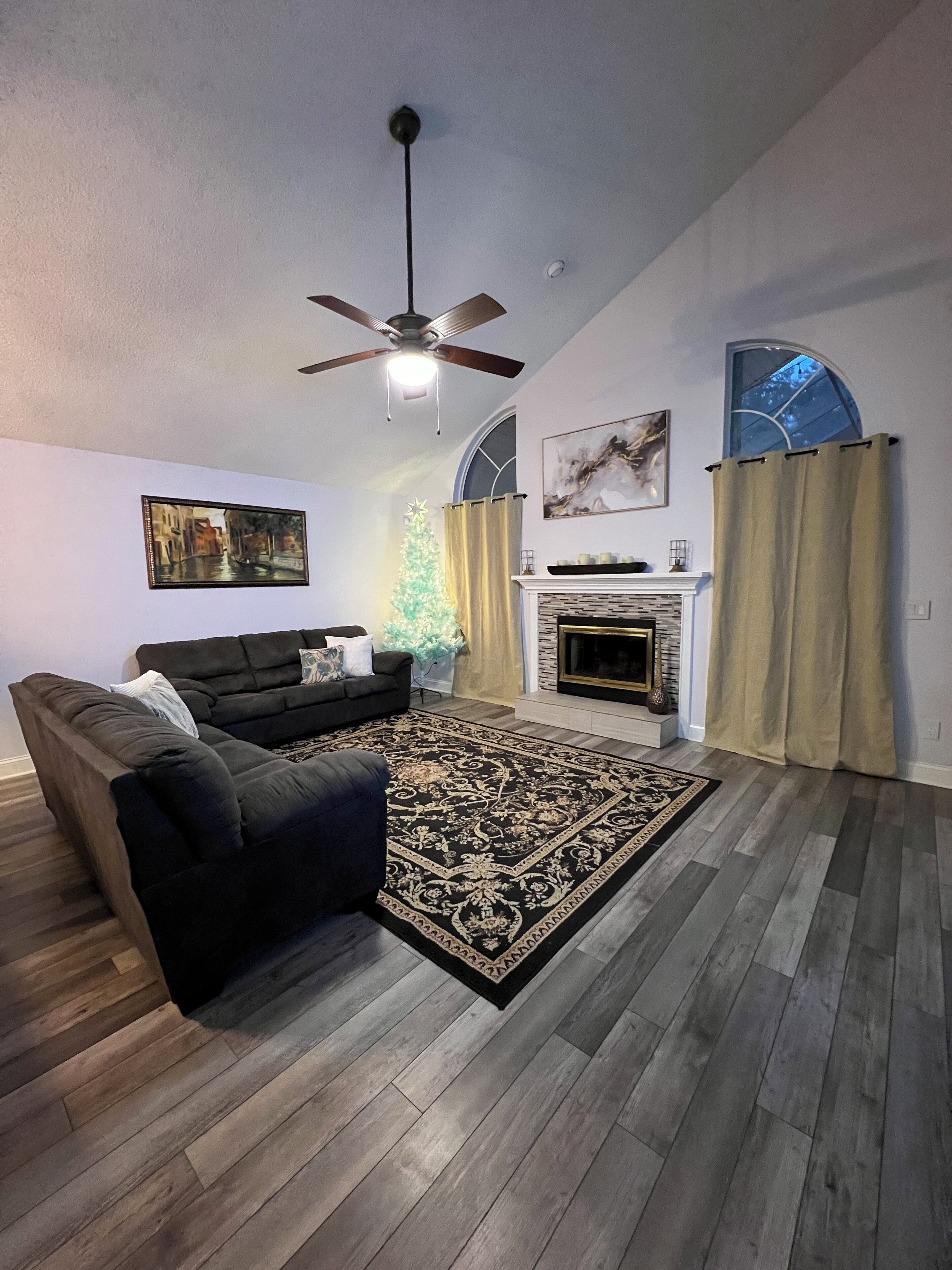 Una sala de estar con un sofá de color oscuro, un tapete estampado, una chimenea y un ventilador de techo.