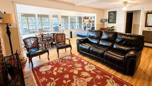 A living room with a leather sofa, a dining area with chairs, a red floral rug, and a ceiling fan. - Hearts Cottage Waterfront Caddo Big Cypress River (Karnack)