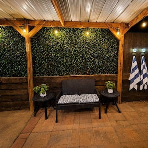 A covered outdoor seating area with a wooden bench, two small tables, and a green leafy wall. - Rest A While Townhome (Tyler)