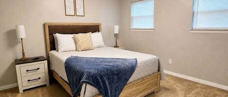Une chambre à coucher avec un lit, des tables de chevet, un luminaire au plafond et deux fenêtres.