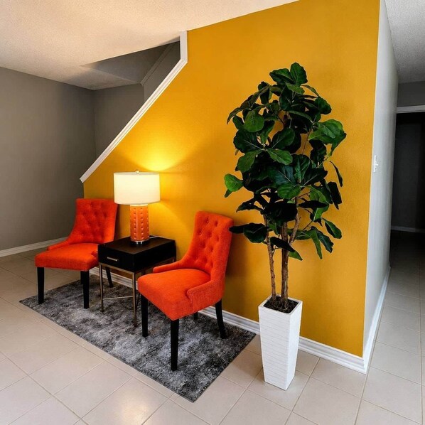 A room with a yellow accent wall, two orange chairs, a small table with a lamp, and a potted plant.