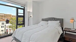 Une chambre moderne avec une grande fenêtre, un lit recouvert de linge de lit blanc, une table de chevet avec une lampe, et une vue sur les bâtiments à l’extérieur.
