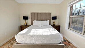 A neatly made bed with a headboard, two bedside tables with lamps, a window with a view, and a patterned rug on the floor. - Warm and Bright Home | Metro | 1 Min Walk | Md (Capitol Heights)