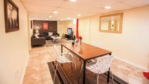 A dining area with a wooden table and white chairs, a sofa, and a TV in the background. - Arlington | Lower Suite | Center | Efficiency (Arlington)