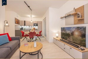 A modern living room with a sofa, a dining table with chairs, a TV, and a kitchen in the background.
