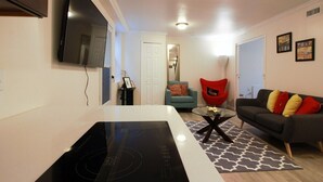 Een moderne woonkamer met een flatscreen televisie, een zwart inductiekookveld, een rode stoel, een grijze bank met kleurrijke kussens, en een kamerkleed met een patroon.