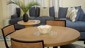 Een woonkamer met een ronde houten tafel, twee stoelen, een bank met opvallende kussens, een kamerplant en een klein bijzettafeltje.