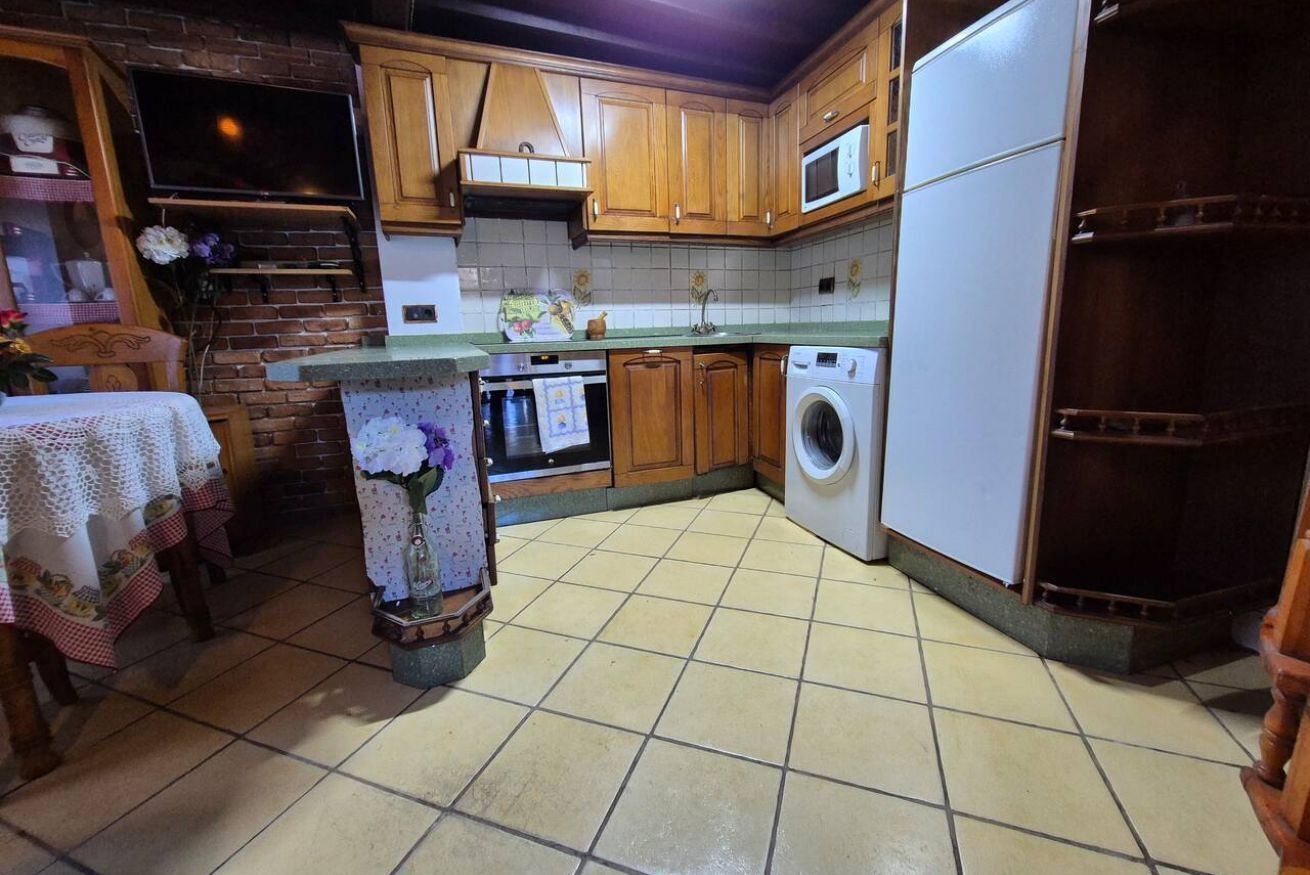 Een keuken met houten kasten, een magnetron, een wasmachine, een betegelde vloer en een televisie aan de muur.