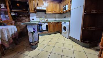 Een keuken met houten kasten, een magnetron, een wasmachine, een betegelde vloer en een televisie aan de muur.