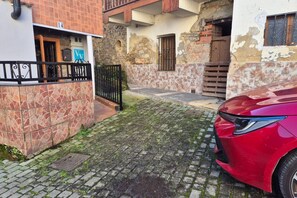 A narrow cobblestone street with a red car parked on the right, a stone wall on the left, and a building with a visible door and window. - 38A01  Casa El Canton (Riosa)