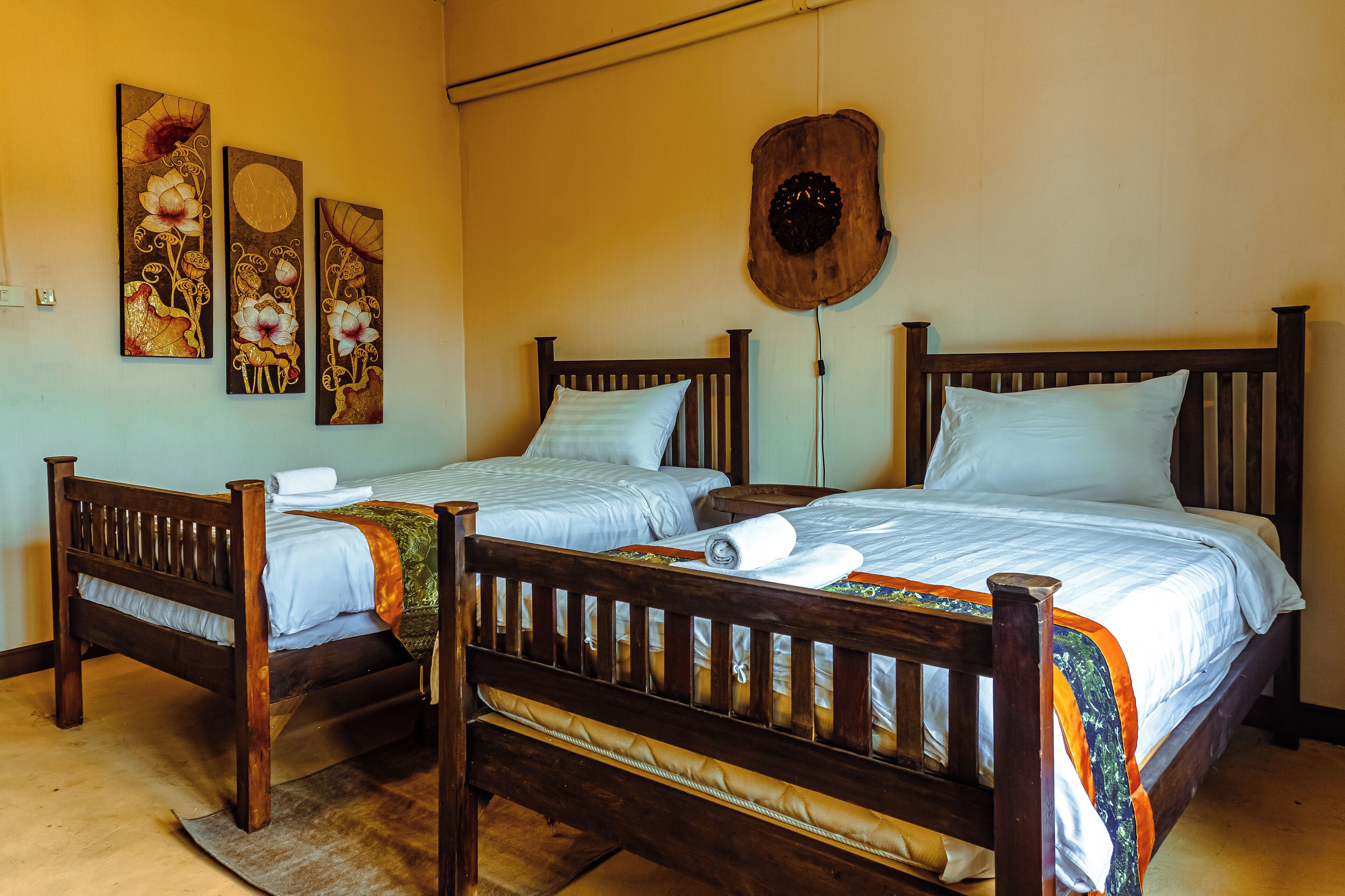 Une chambre avec deux lits en bois, chacun muni d’un oreiller et d’une serviette enroulée. Des œuvres d’art sont accrochées aux murs, au-dessus des lits.