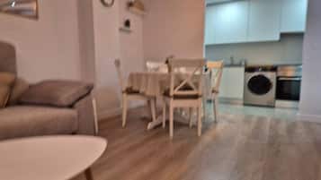 Una sala de estar con un sofá, una mesa redonda y un comedor con sillas. Se observa una cocina con armarios y electrodomésticos al fondo.