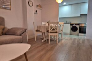 A living room with a sofa, a round table, and a dining area with chairs. There is a kitchen with cabinets and appliances in the background. - 650A Apartamento moderno, acogedor y con garaje (Oviedo)