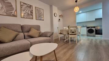 Una sala moderna con un sofá, una mesita y un comedor con sillas. Se aprecia una cocina con armarios y electrodomésticos al fondo.