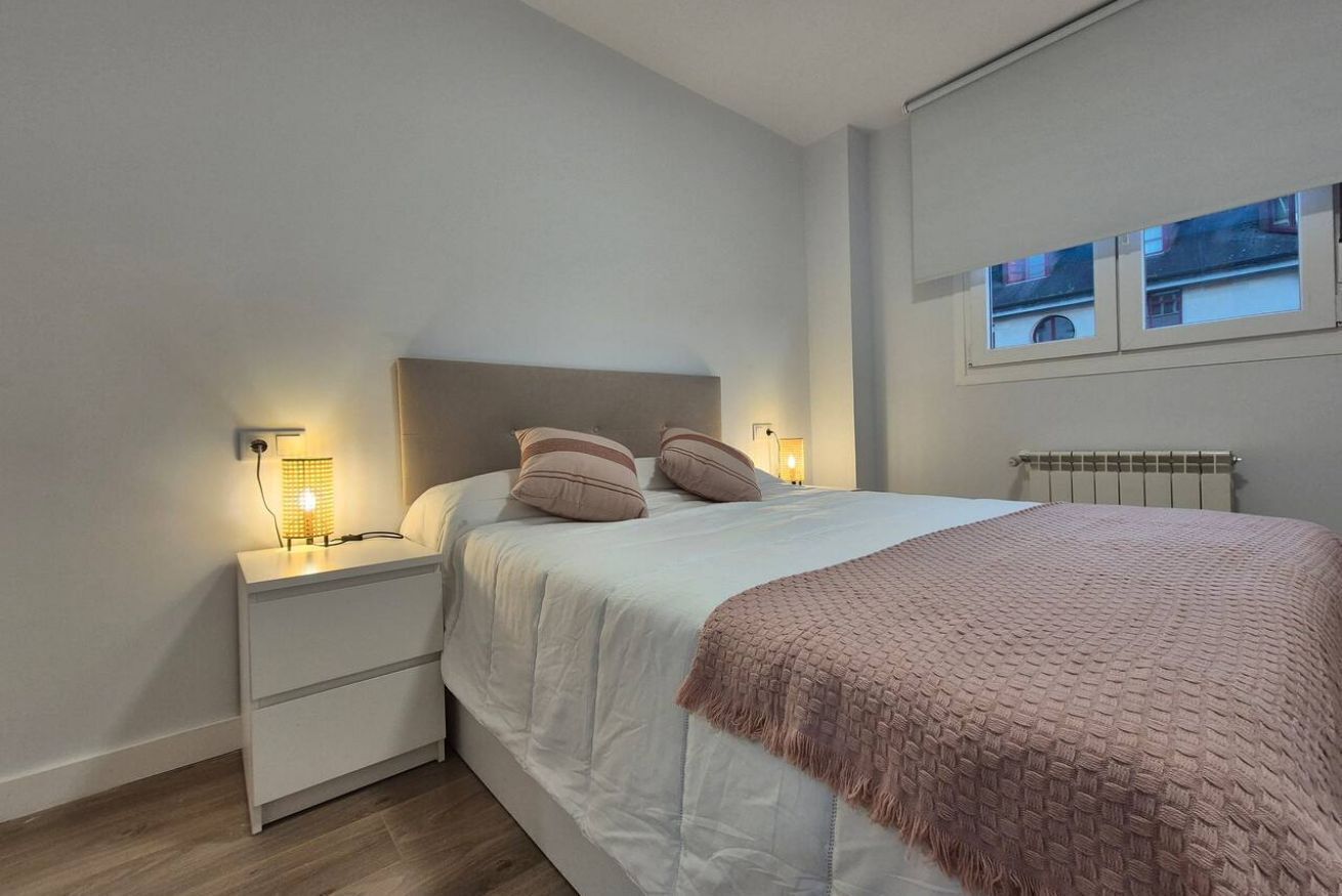 Un dormitorio con cama, lámparas de noche, mesita de noche, ventana y radiador.