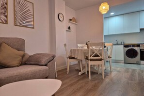 A living room with a sofa, a dining table with chairs, a clock, and a washing machine in the background. - 650A Apartamento moderno, acogedor y con garaje (Oviedo)