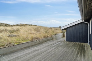 A wooden deck with a metal railing, a dark building with a window, and a grassy hill in the background.