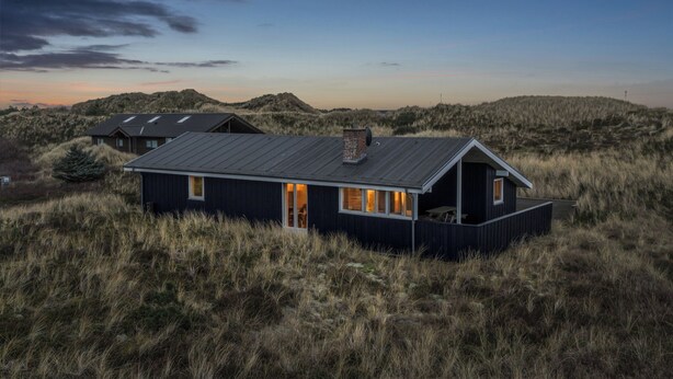 A modern house with a flat roof, large windows, and a balcony, surrounded by tall grass and hills.