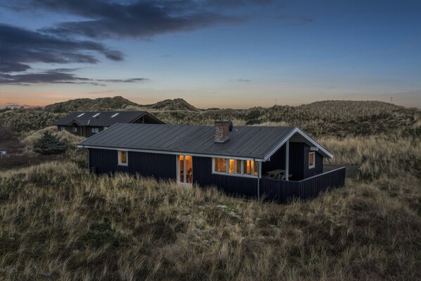 A modern house with a flat roof, large windows, and a balcony, surrounded by tall grass and hills.