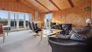 A spacious room with a wooden ceiling, a leather sofa, a dining table, and a large sliding glass door leading to an outdoor area.