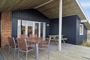 A covered outdoor area with a wooden deck, a brick wall, and a dark-colored building with a glass door and windows.