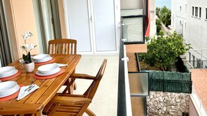 Ein Holztisch mit weißen Tellern und einer Vase mit weißen Blumen auf einem Balkon mit Blick auf das Meer und angrenzende Gebäude.